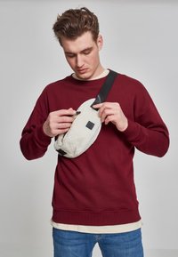 Fanny pack i kremfarget fleece med svart bunn, oval form, justerbar stropp og glidelåslomme. Modell iført en burgunder genser.