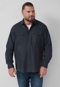 Donkergrijze overhemd met knopen, twee borstzakken, lange mouwen en een klassieke kraag. Draagt over een wit T-shirt en gecombineerd met blauwe jeans.