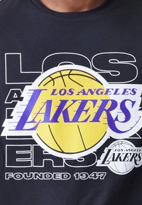 Čierne tričko s grafickým dizajnom, na ktorom je žltý basketbal a fialový text "LOS ANGELES LAKERS" prekrývajúci biele kontúry.