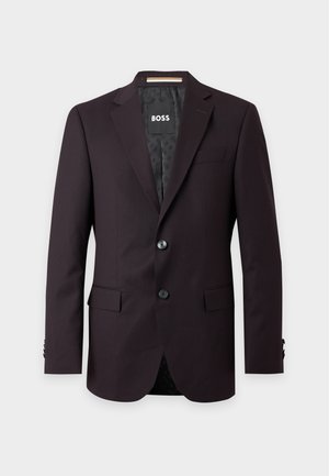 Un homme porte un costume sombre deux pièces avec une chemise blanche et des mocassins noirs sur un fond clair uni.
