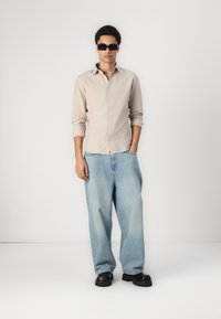 Light beige button-up shirt, relaxed-fit light blue jeans, and chunky black footwear. Model wears dark sunglasses. Simple background.
