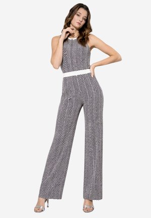 Frau posiert in einem ärmellosen Jumpsuit mit schwarz-weißem Chevron-Muster, weißem Taillenbund und silbernen High-Heel-Sandalen vor weißem Hintergrund.