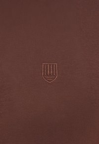 Καφέ ύφασμα με ένα μικρό κεντητό λογότυπο σε σχήμα ασπίδας με κάθετες ρίγες στο κέντρο.