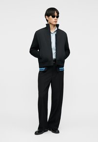 Άνδρας με μαύρο μπουφάν bomber πάνω από ριγέ πουκάμισο, μαύρο παντελόνι με ευρύ πόδι, μαύρα δερμάτινα παπούτσια και μαύρα γυαλιά ηλίου, στέκεται με τα χέρια στις τσέπες.