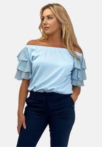 Jasnoniebieska bluzka typu off-the-shoulder z warstwowymi rękawami z falbanami, gładki materiał, w połączeniu z ciemnymi spodniami z zapięciem na dwa guziki.