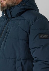 Granatowa pikowana kurtka puchowa z wysokim kołnierzem, zamkiem błyskawicznym i przednią kieszenią. Na ramieniu znajduje się czarna materiałowa naszywka.