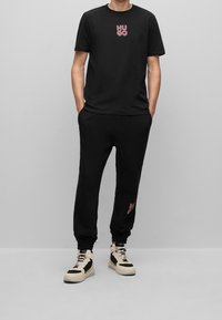 Sort t-shirt med pink "HUGO" logo, sorte sweatpants og beige og sorte højhælede sneakers med snørebånd. Glat stof, afslappet pasform.