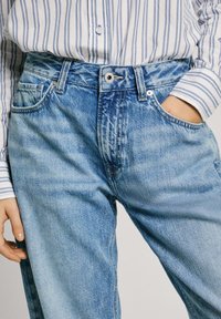Bledě modré džíny z denimu s vysokým pasem, se stříbrným knoflíkem a zipem, a bočními kapsami. Materiál vypadá měkký a texturovaný.