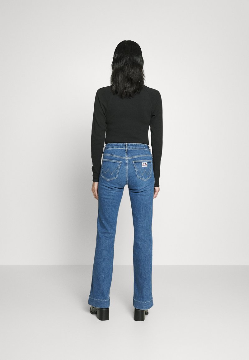 Person steht mit dem Rücken zur Kamera, trägt ein schwarzes Langarmshirt, eine blaue Jeans mit Hintertaschen und schwarze Absatzschuhe vor einem weißen Hintergrund.