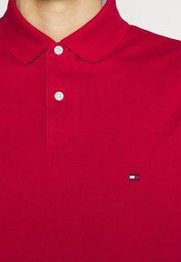 Punainen puuvilla-pikeepaita, jossa on klassinen kaulus, kaksi valkoista nappia ja pieni logo sinisessä, valkoisessa ja punaisessa vasemmassa rinnassa.