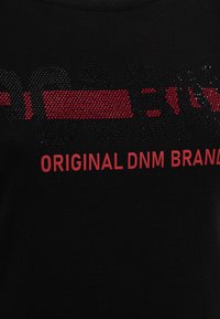 Černé tričko s kulatým výstřihem, které je zdobené vyvýšenými černými a červenými kamínky s nápisem "ORIGINAL DNM BRAND." Měkký bavlněný materiál, ležérní design.