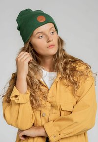 Zielona, prążkowana dzianinowa czapka z brązową, okrągłą skórzaną łatką, noszona z żółtą kurtką dżinsową na białej koszuli, z długimi, falującymi włosami.