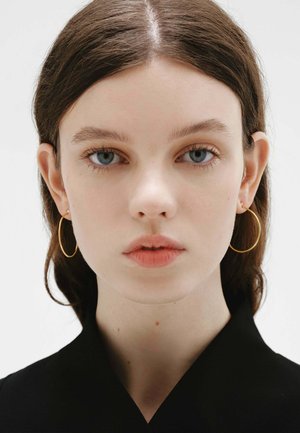 Joven con piel clara y ojos azules, lleva grandes pendientes de aro dorados y una camiseta negra, mirando directamente a la cámara contra un fondo blanco.