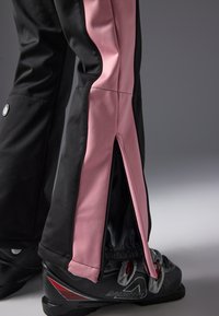 Zwarte skibroek met roze zijpanele, met een ritssluiting bij de enkels. Materiaal lijkt glad en aansluitend. Zwarte skibotten zichtbaar.