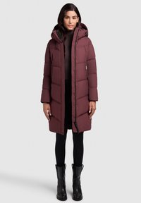 Bordeaux gewatteerde lange jas met een capuchon, voorzien van een ritsluiting, zijzakken en een gestructureerde stof. Gedragen over zwarte leggings en laarzen.