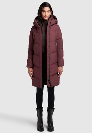 Bordeaux gewatteerde lange jas met een capuchon, voorzien van een ritsluiting, zijzakken en een gestructureerde stof. Gedragen over zwarte leggings en laarzen.