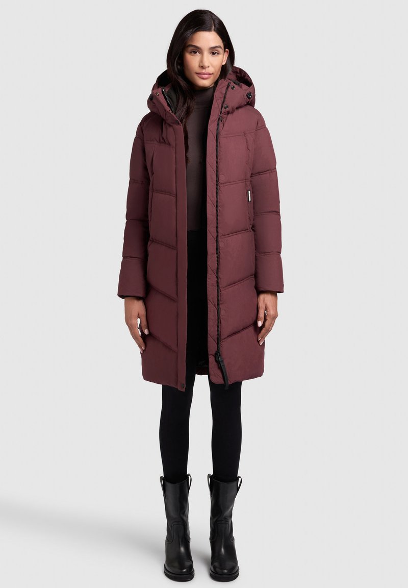 Bordeaux gewatteerde lange jas met een capuchon, voorzien van een ritsluiting, zijzakken en een gestructureerde stof. Gedragen over zwarte leggings en laarzen.