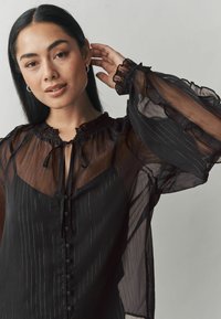 Blusa nera trasparente con righe verticali, colletto arruffato e maniche a sbuffo. Presenta una chiusura con laccetto e dettagli con bottoni lungo la parte anteriore.