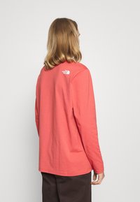 Pitkähihainen coral-värinen puuvillapaita, jossa on pyöreä kaula-aukko, suora helma ja pieni valkoinen logo takana. Pehmeä pinta.