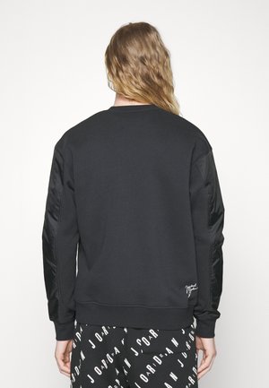 Personne aux longs cheveux ondulés porte un sweat-shirt noir avec des manches matelassées et un pantalon noir orné du texte blanc « Jordan », vue de dos.
