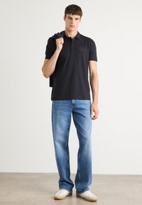 Camisa polo azul-marinho de mangas curtas com um logótipo de crachá, combinada com jeans de perna reta azul-claro e ténis brancos, suéter a cair sobre o ombro.