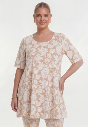 Lächelnde Frau mit zurückgebundenen Haaren trägt eine beige Tunika und Hose mit weißem Blumenmuster, Creolen und ein goldenes Armband, steht vor weißem Hintergrund.