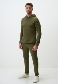 Felpa verde oliva con pantaloni coordinati in materiale morbido e elasticizzato. Presenta una tasca a marsupio, polsini a costine e un design minimalista.