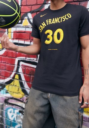 Person trägt ein schwarzes San Francisco 30 Shirt und dreht einen schwarz-gelben Basketball mit einem Finger vor einer Graffiti-Wand.