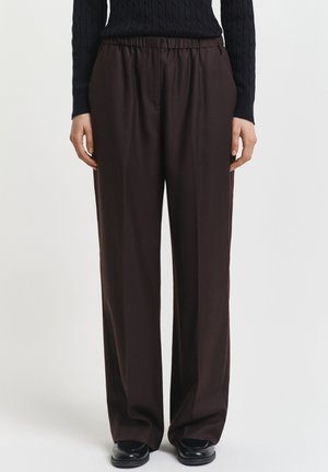 Person trägt locker sitzende dunkelbraune Hosen mit elastischem Bund und schwarzen Schuhen, kombiniert mit einem schwarzen, langärmligen Strickoberteil, steht still.