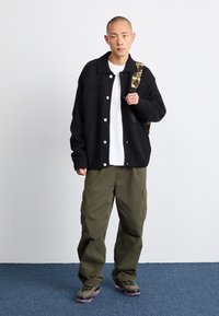 Cardigan nero a maglia con grandi bottoni, indossato sopra una T-shirt bianca, abbinato a pantaloni cargo verde oliva e uno zaino fantasia.