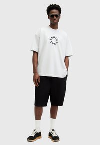 Hvit oversized t-skjorte med sirkulær svart grafisk tekst, paired med svarte shorts, hvite sokker og svarte joggesko med gummisåle.