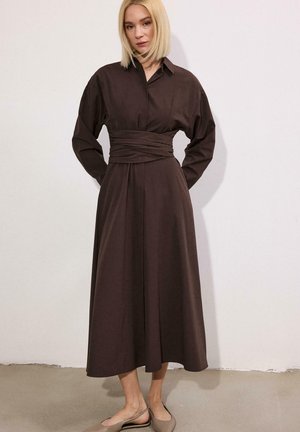 Blondínka v dlhých rukávoch, tmavohnedých midi šatách so širokým pásom, stojaca bosá pred bielou stenou.