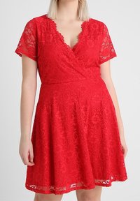 Kvinna klädd i en kortärmad klänning i röd spets med en vågig V-ringning och utsvängd kjol.