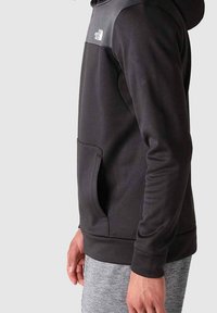 Sweat à capuche noir avec un panneau gris à l'épaule, doté d'une poche avant et de poignets côtelés, fabriqué en tissu doux.