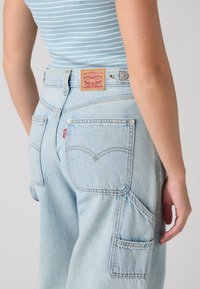 Άτομο που φοράει ανοιχτό μπλε τζιν Levi's με πίσω τσέπες και ριγέ μπλε και λευκή αμάνικη μπλούζα.
