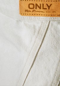 Vaaleansininen farkkukangas, jonka pinnassa on rakenne, ja jossa on ruskea nahkaleima, johon on kohokuvioitu "ONLY Blue Denim anno 1995."