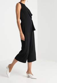 Czarny kombinezon z dopasowanym korpusem, rozszerzanymi nogawkami i wiązaniem w talii. Noszony z białymi klapkami na słupku.