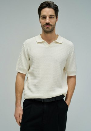 Hombre con cabello oscuro y barba, lleva un suéter de manga corta color crema con textura y pantalones negros, de pie con una mano en el bolsillo.