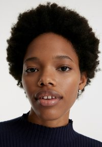 Nahaufnahme einer Person mit kurzen, strukturierten schwarzen Haaren, die einen marineblauen Ripppulli und kleine silberne Ohrhänger trägt. Natürlicher Makeup-Look, weiche Haut.