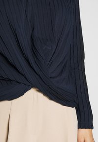 Marinblå långärmad topp med en strukturerad ribbad design, med en tvistad frontdetalj. I kombination med beige midjehöga byxor.