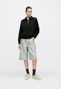 Μαύρο φούτερ με φερμουάρ και γιακά, σε συνδυασμό με μεταλλικό ασημί σορτς. Το μοντέλο φοράει μαύρες κάλτσες και ανοιχτό μπεζ αθλητικά παπούτσια.