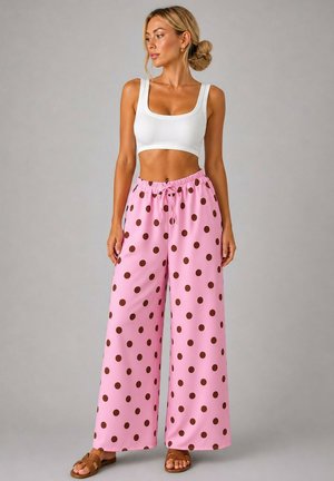 Femme portant un débardeur blanc court, un pantalon large rose à pois marron et des sandales marron, debout devant un fond gris.