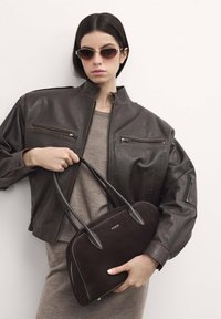 Giacca bomber in pelle marrone con tasche con zip, abbinata a un vestito in maglia taupe. Il modello tiene in mano una borsa in suede marrone scuro con logo.