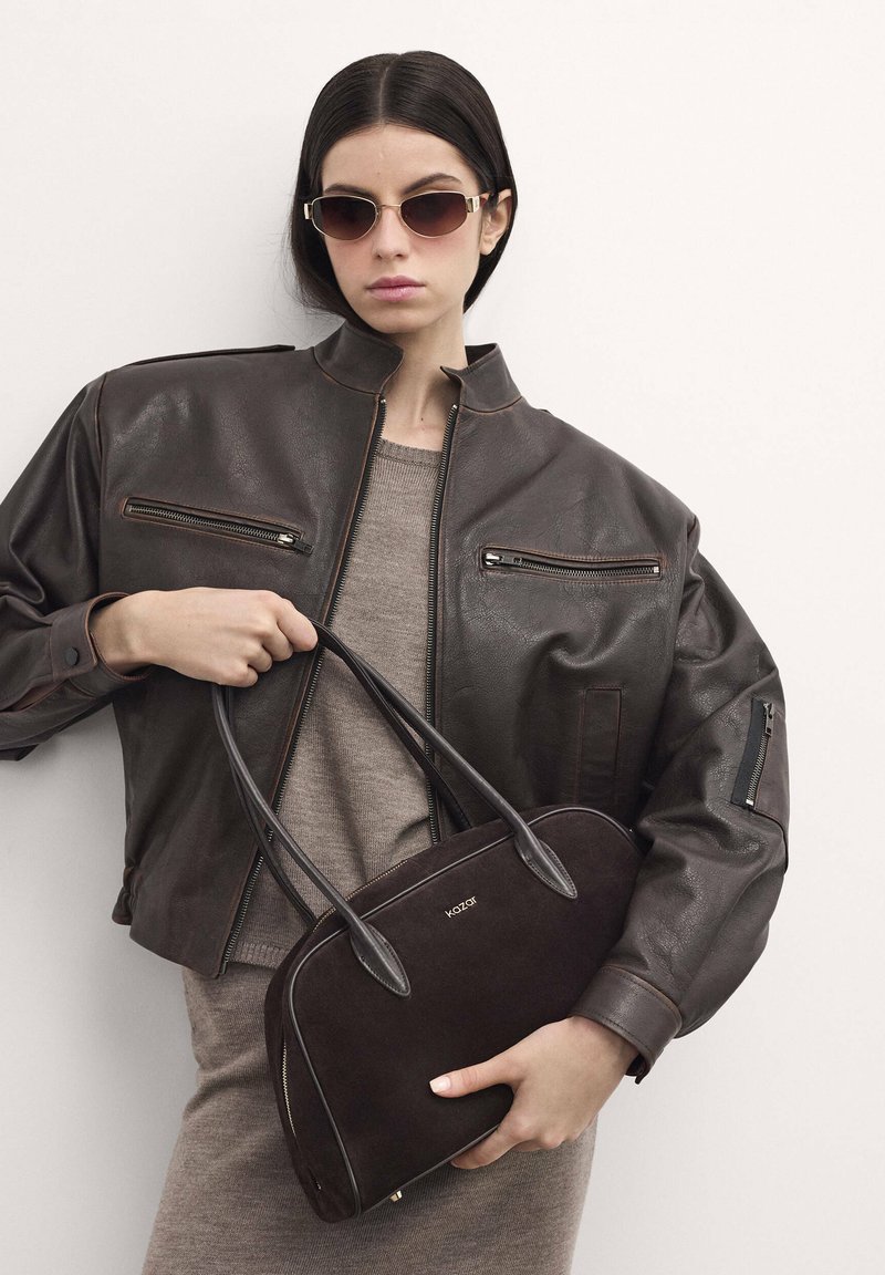 Giacca bomber in pelle marrone con tasche con zip, abbinata a un vestito in maglia taupe. Il modello tiene in mano una borsa in suede marrone scuro con logo.