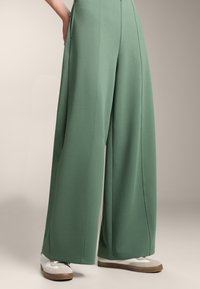 Zelene široke hlače izdelane iz mehkega blaga, z visokim pasom, stranskimi žepe in detajli šivanja. Skombinirane z belimi in sivimi športnimi copati.