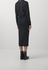 Vrouw in een zwarte lange mouwtop en bijpassende midi-jurk met witte stippen, gecombineerd met zwarte enkellaarsjes, met de achterkant naar de camera gekeerd.