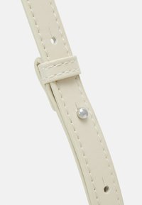 Kožený řemínek hodinek v krémově bílé barvě s bílým prošitím, dírkami pro nastavení velikosti a stříbrným zdobením poblíž oblasti přezky.