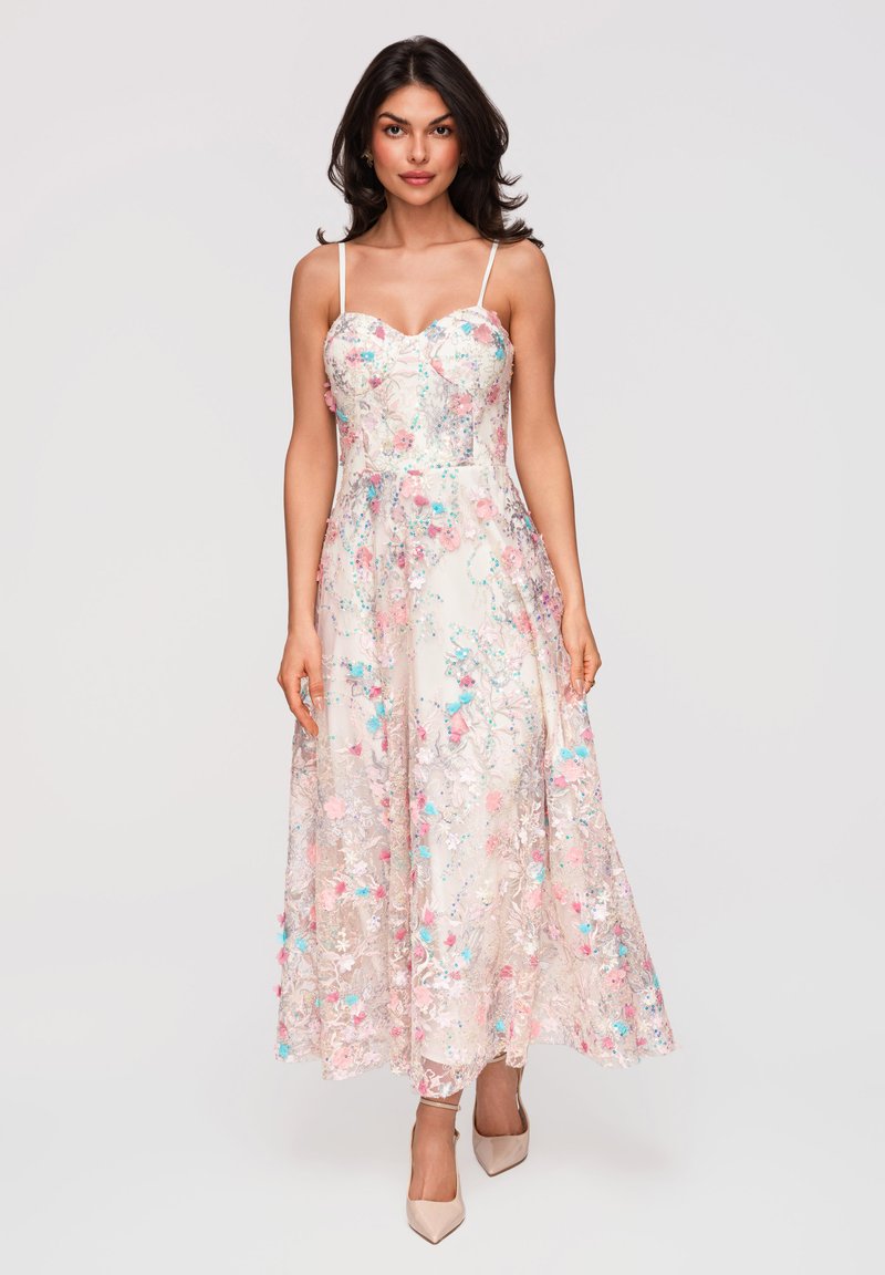 Femme portant une robe longue fleurie sans manches avec des broderies roses, bleues et blanches, associée à des chaussures à talons hauts couleur nude.