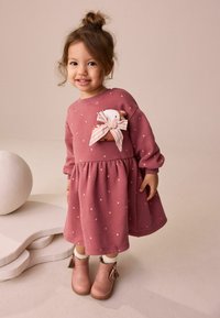 Roze jurk met stippen, lange mouwen en een afgeronde onderkant. Heeft een dierenapplicatie met een gestreepte strik. Bruine enkellaarsjes met franjes.