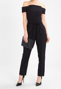 Kvinna iklädd en svart off-shoulder jumpsuit med midjeband, håller en svart clutchtvätt och bär svarta högklackade skor.